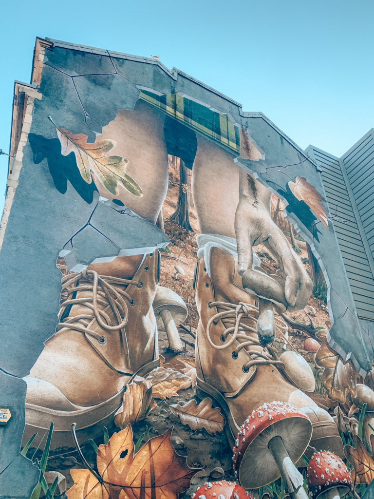 This mural is painted to appear as if the wall is cracked, and from it emerges a leafy forest, with two large boots and a hand reaching for a mushroom