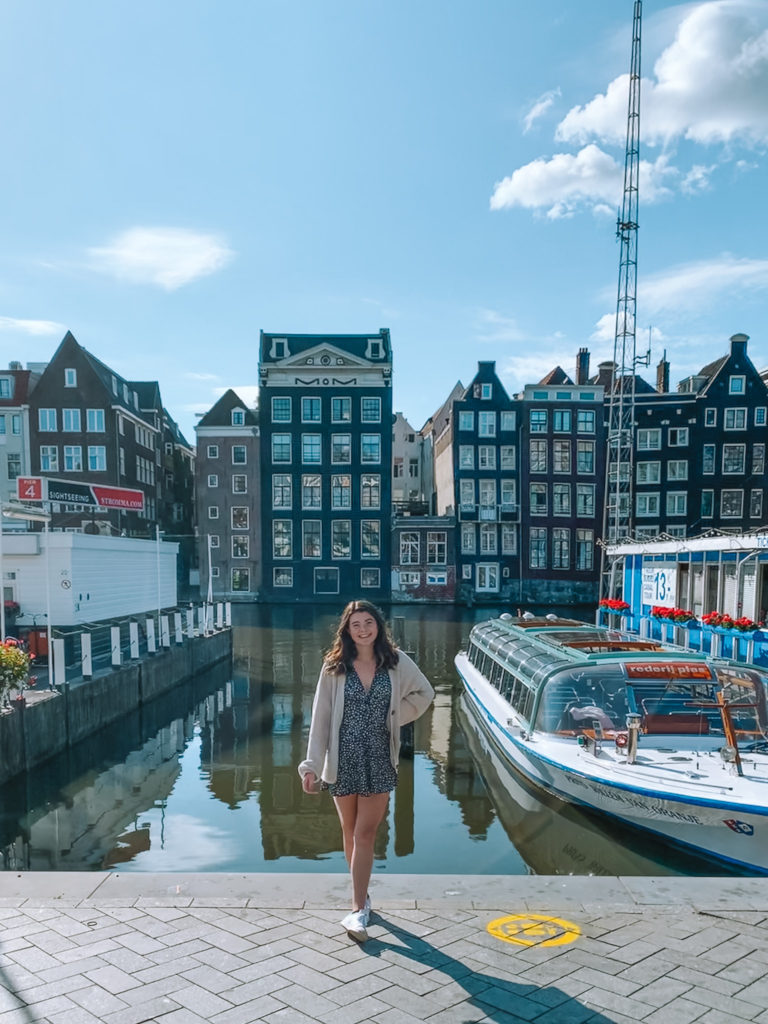 a girl stands in front of some canal houses and boats at Damrak
