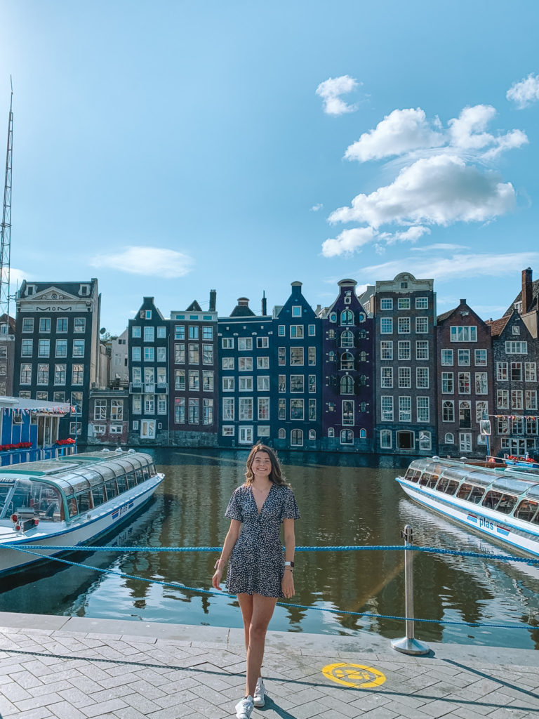 a girl stands in front of some canal houses and boats at Damrak