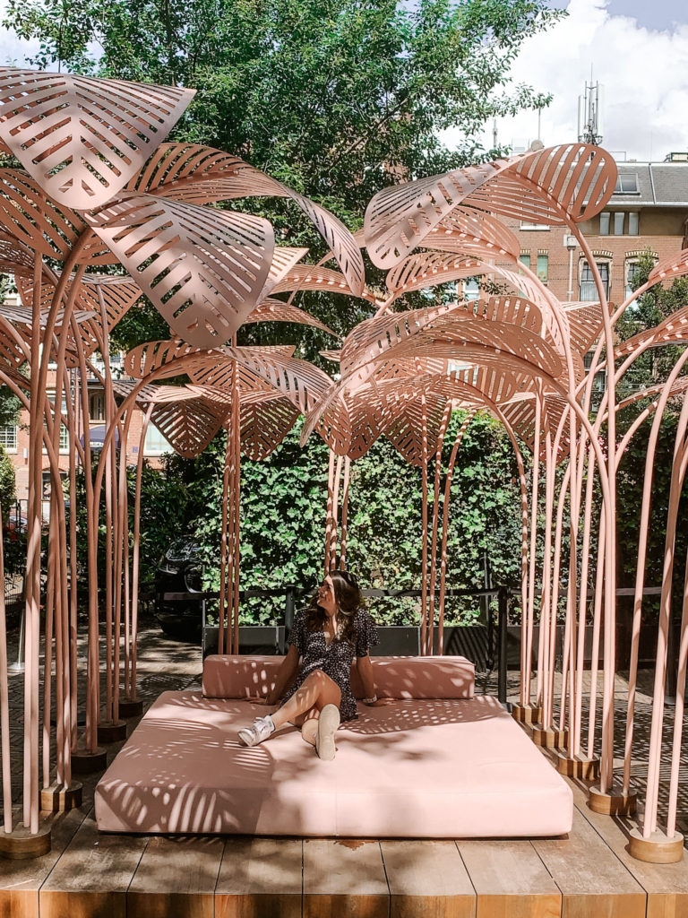 a girl sits on a bed with pink palm trees surrounding it, at the moco museum in Amsterdam
