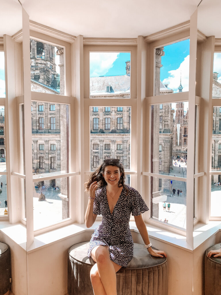 a girl smiles sitting in a window looking out to Dam Square in Amsterdam