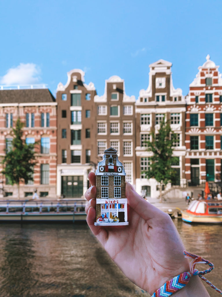 a hand holds a canal house magnet in front of some canal houses in Amsterdam