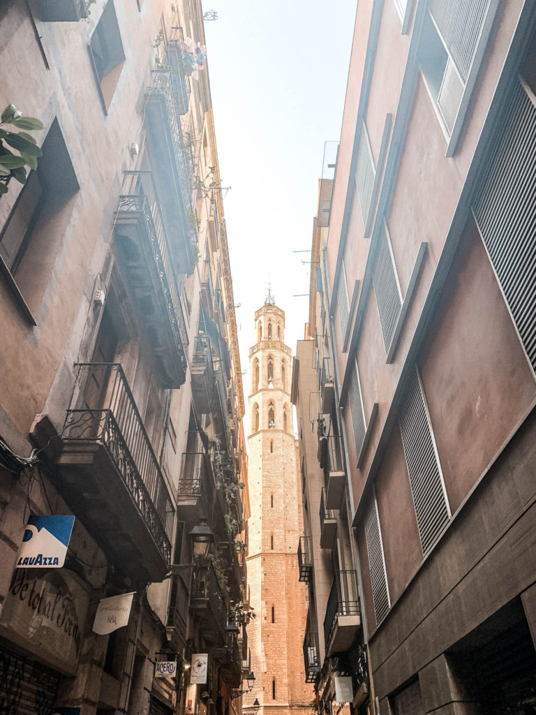 a street in the el born neighbourhood of Barcelona with a tower of a Santa Maria del Mar basilica visible at the end