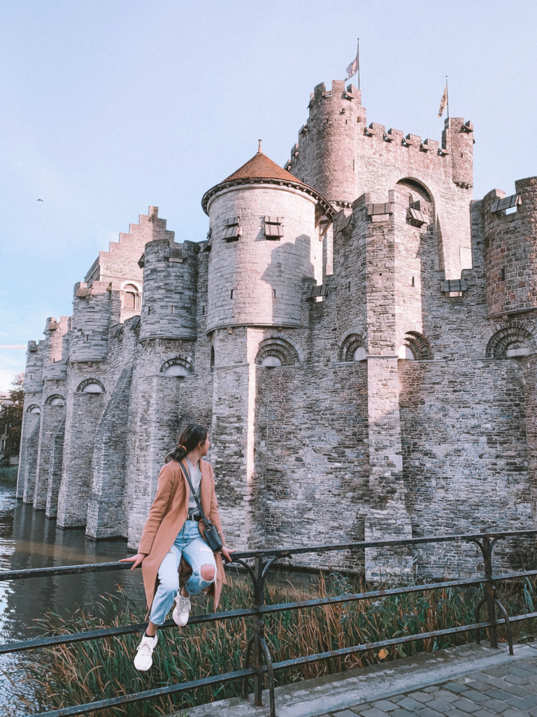 A girl sits on a railing and looks back at Gravensteen castle and its moat in Ghent