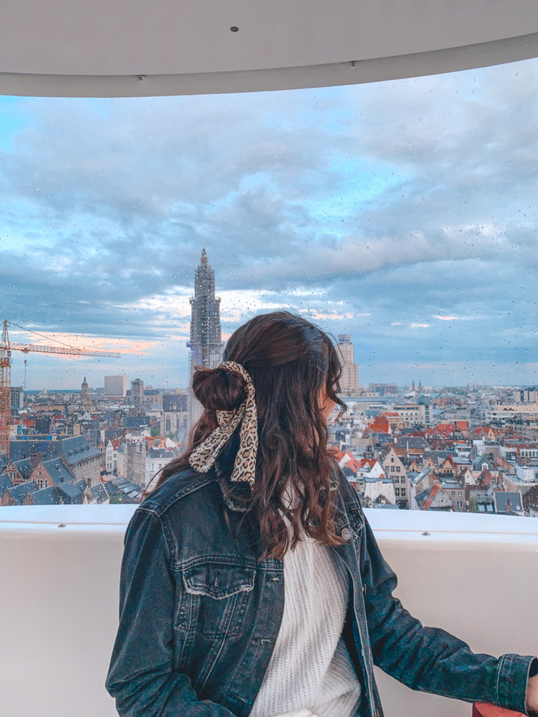 A girl sits in a Ferris Wheel car and looks behind her over a view of Antwerp from above