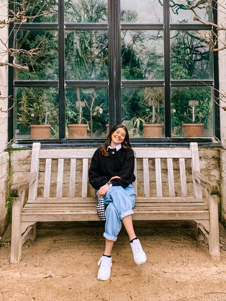 A girl sits on a bench in front of a greenhouse window at Kruidtuin, Leuven