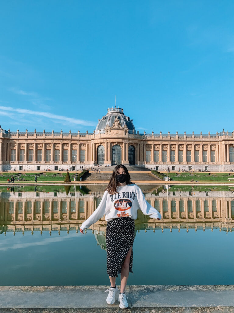 a girl in a black skirt and graphic sweatshirt stands in front of the royal museum for central Africa, with a perfect reflection in the pond in front