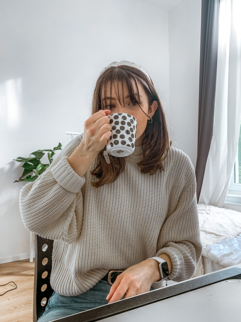 a girl in a grey knitted jumper drinks from a polka dot mug in her bedroom